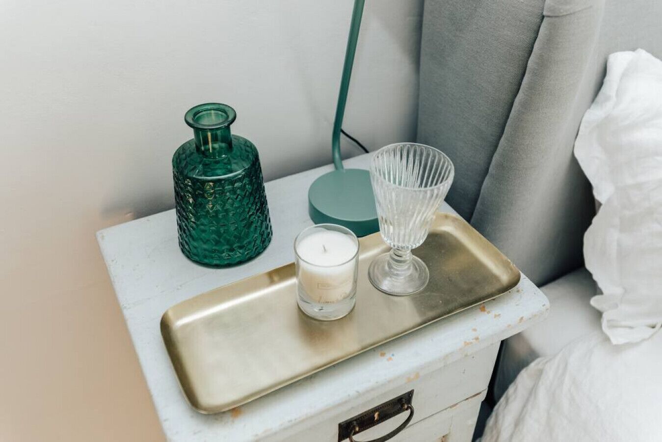 Una mesita de noche con un cajón blanco, una botella de vidrio verde, una vela de vidrio transparente y un candelabro de vidrio.