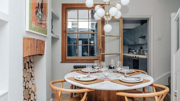 Een eethoek met een ronde tafel voor zes personen, een houten stoel, een moderne kroonluchter, een schouw met bakstenen en een raam met uitzicht op de stad.