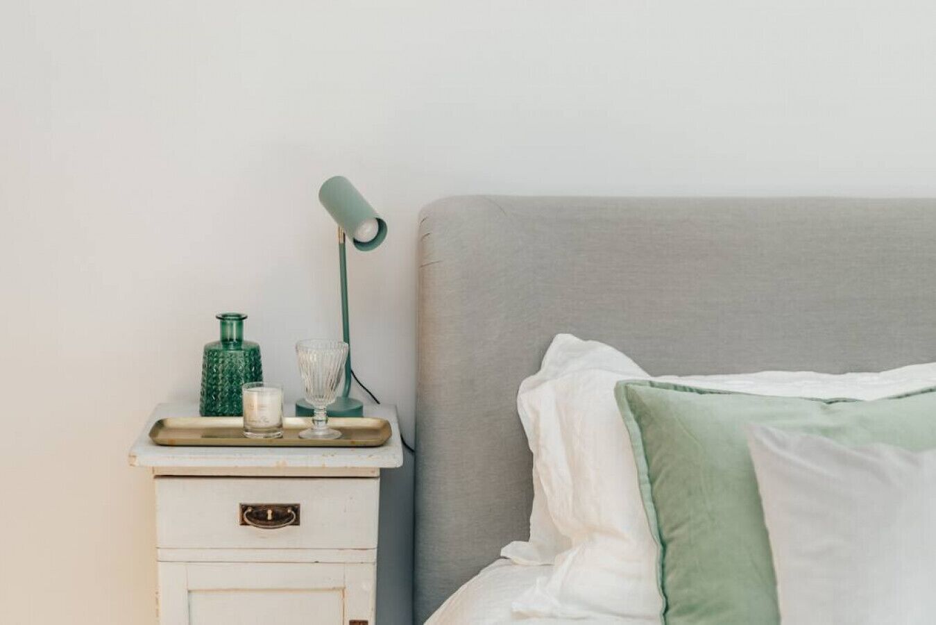 Una cama bien hecha con almohadas blancas y verdes, una mesita de noche con una lámpara, un vaso y una botella verde.