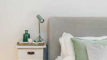 Una cama bien hecha con almohadas blancas y verdes, una mesita de noche con una lámpara, un vaso y una botella verde.