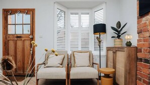 Een kamer met twee beige fauteuils, een houten kast, een vloerlamp en een raam met witte luiken.
