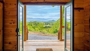 Une cabane en bois dotée d’une grande porte ouverte donnant sur une terrasse offrant une vue panoramique sur une forêt et des collines.