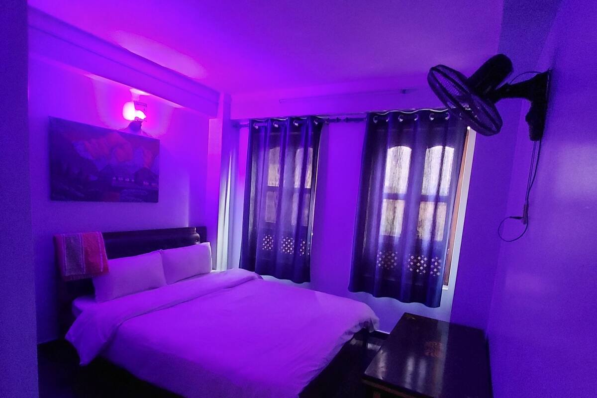 Een kamer met een bed, een bureau, een ventilator aan de muur en gordijnen.