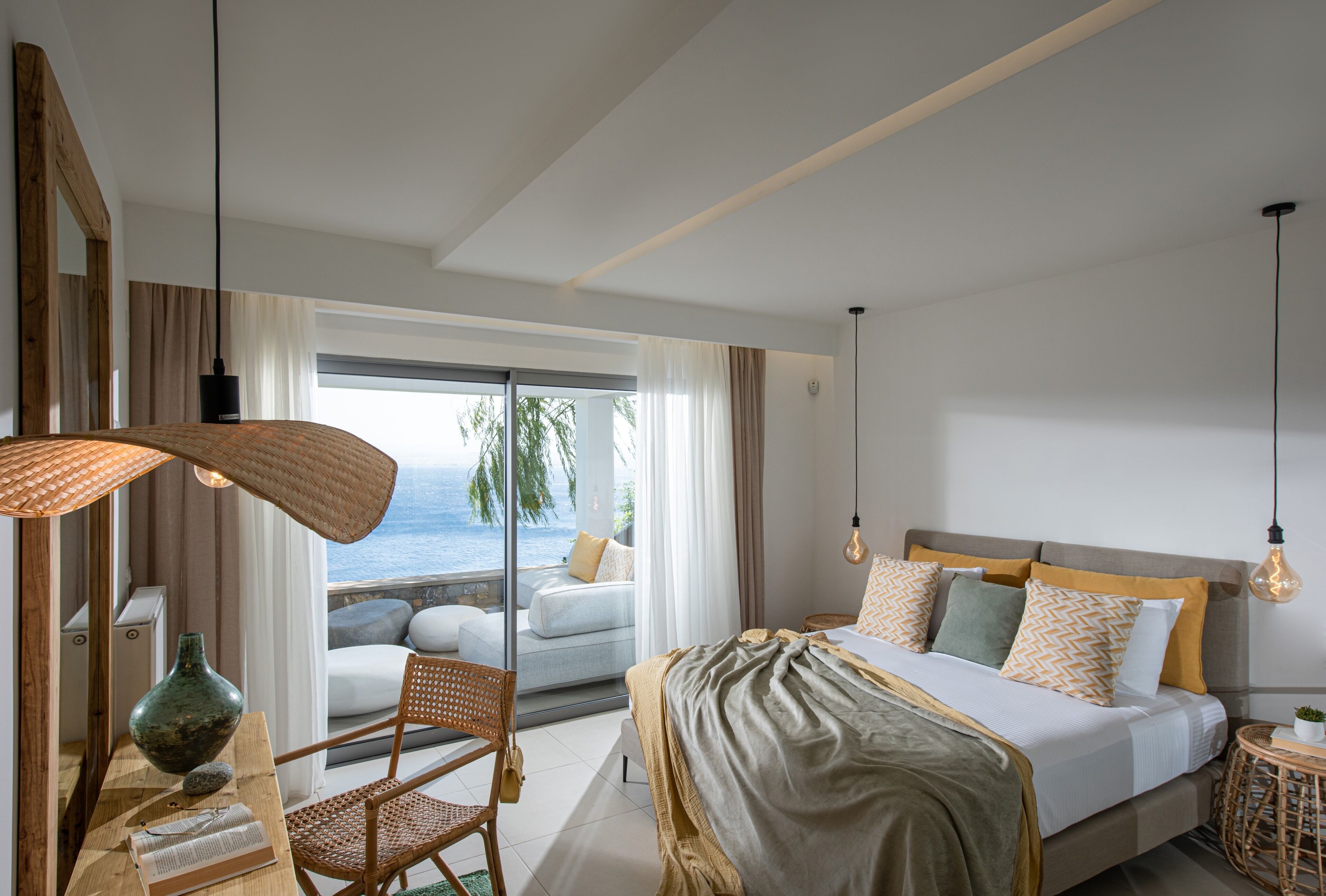 A modern bedroom with a large bed, a view of the ocean, a wicker chair, and a wooden nightstand.