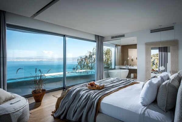 A modern bedroom with a large window, a bed, a bedside table, and a view of the sea.
