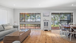 A modern living room with a sofa, coffee table, and dining area with chairs. Large windows provide views of the outdoors. - Kullaroo Lakehouse - Waterfront Reserve (Summerland Point)