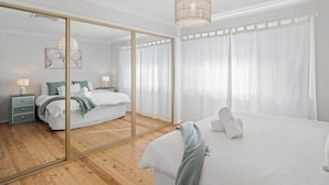 A bedroom with a large mirror, a bed with white linens, a bedside table with a lamp, and a hanging light fixture. - Kullaroo Lakehouse - Waterfront Reserve (Summerland Point)