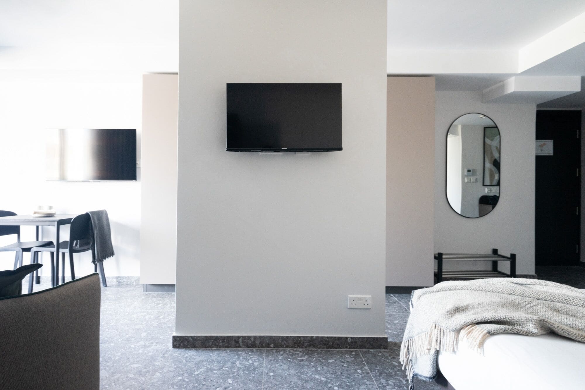 A modern hotel room with a flat-screen TV mounted on the wall, a large mirror, a bed with a white blanket, and a seating area with chairs and a table.