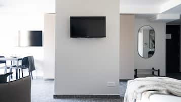 A modern hotel room with a flat-screen TV mounted on the wall, a large mirror, a bed with a white blanket, and a seating area with chairs and a table.