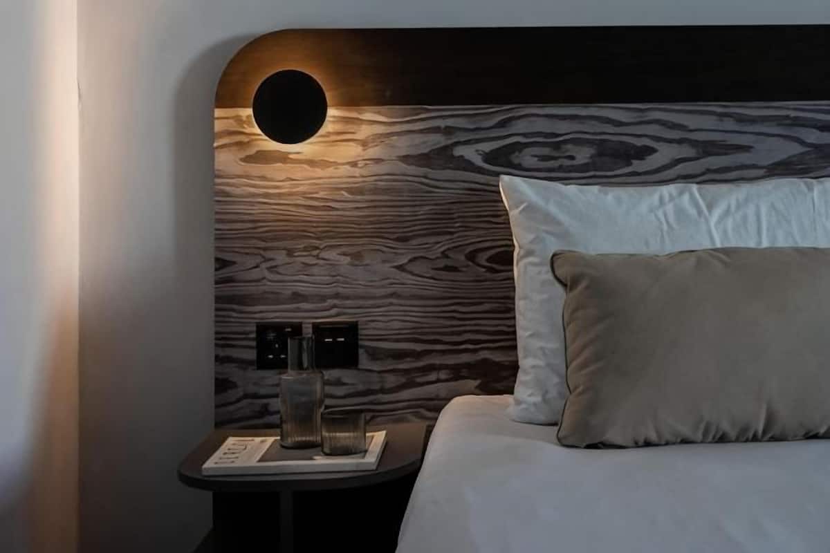 Una camera da letto con una luce circolare a parete, un comodino con una lampada e un letto con un cuscino.