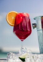 A glass of red beverage with an orange slice garnish, ice cubes, and a lime wedge, placed on a table with a metal pitcher in the background.