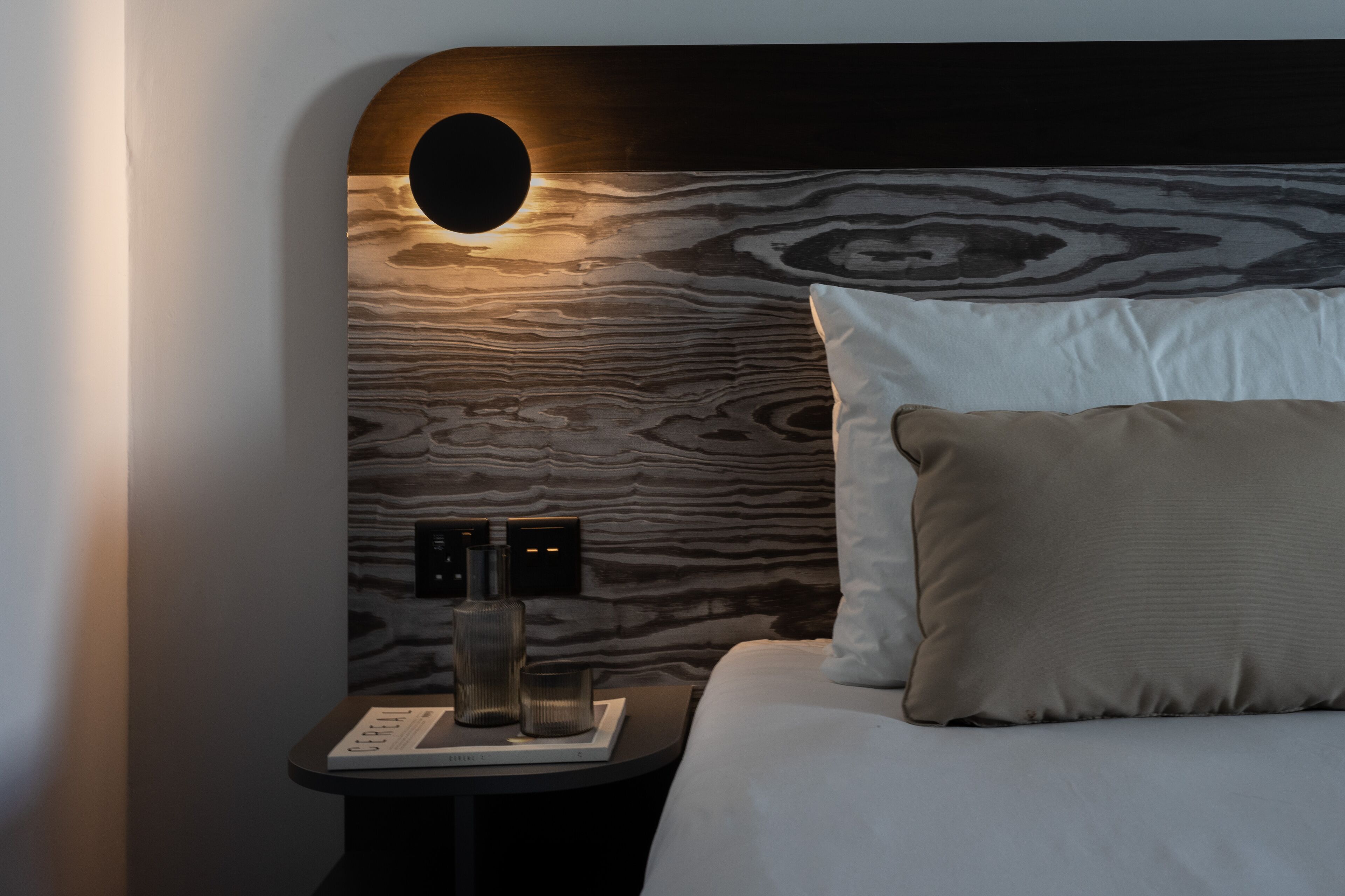 Una habitación de hotel moderna con un cabecero de madera, una mesita de noche con lámpara y una cama bien tendida con almohadones.