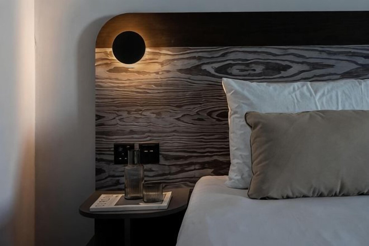 Une chambre avec un luminaire mural circulaire, une table de chevet avec une lampe et un lit avec un oreiller.