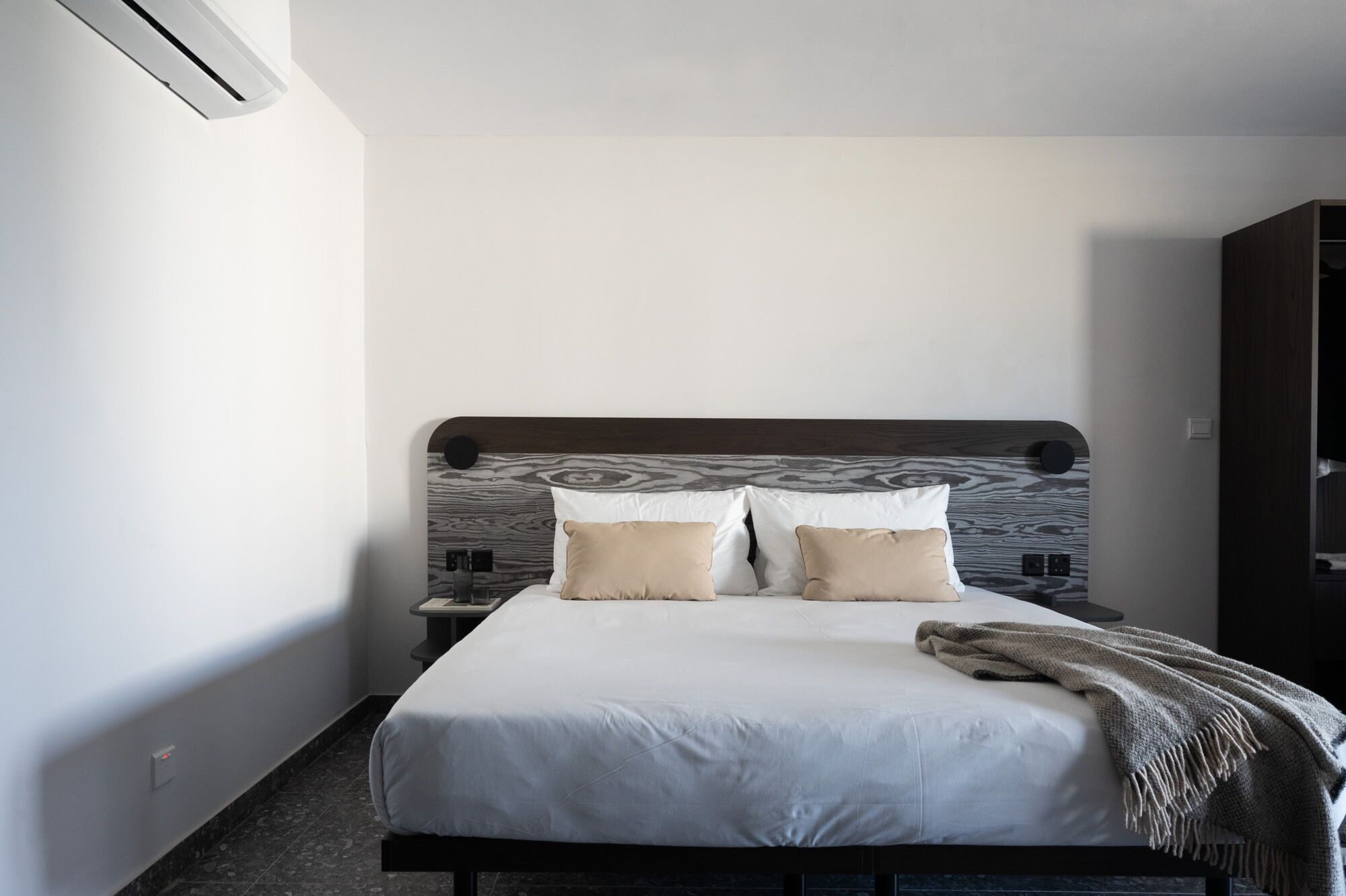 Una cama bien hecha con tablero de madera, dos almohadas, una colcha y una mesita de noche con una lámpara.