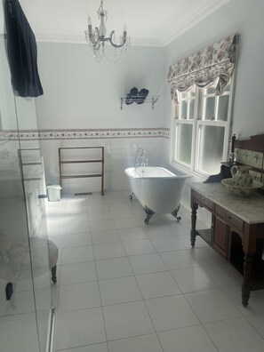 Un baño con una bañera independiente, un tocador de madera, una lámpara de araña y un ventanal con cortinas florales.