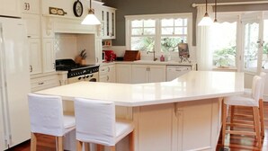 A kitchen with white cabinetry, a central island with bar stools, a stove, and a window with a view of greenery. - Large Cottage - Heart of Olinda - Old Police station (Olinda)