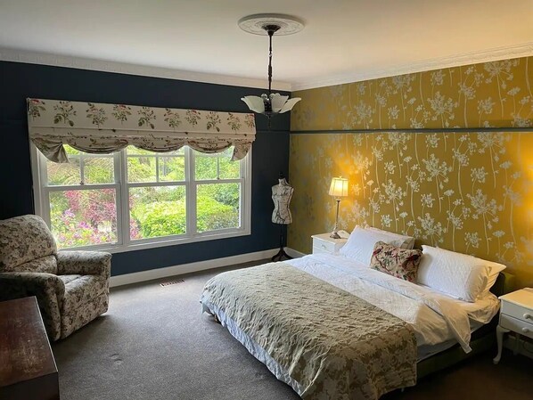 Un dormitorio con una cama grande, una silla, un aparador y una ventana con cortinas florales.