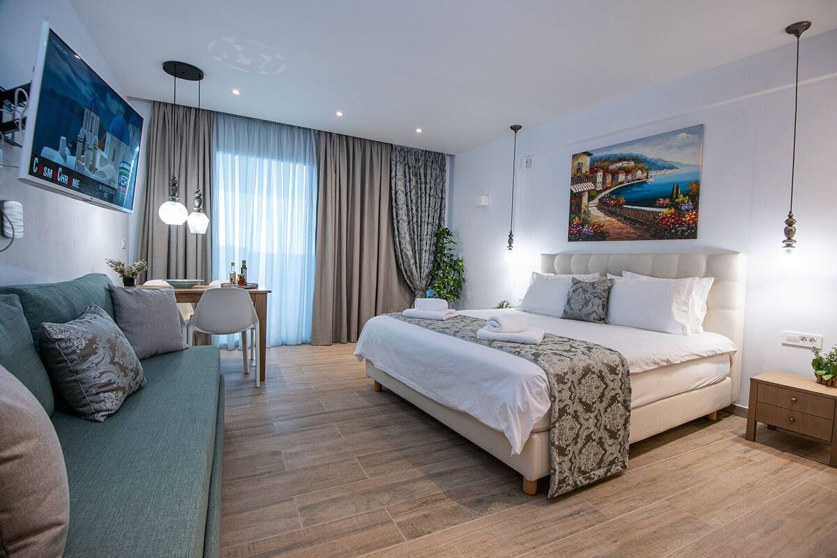 A modern hotel room with a large bed, a sofa, a desk, and a painting of a coastal scene.
