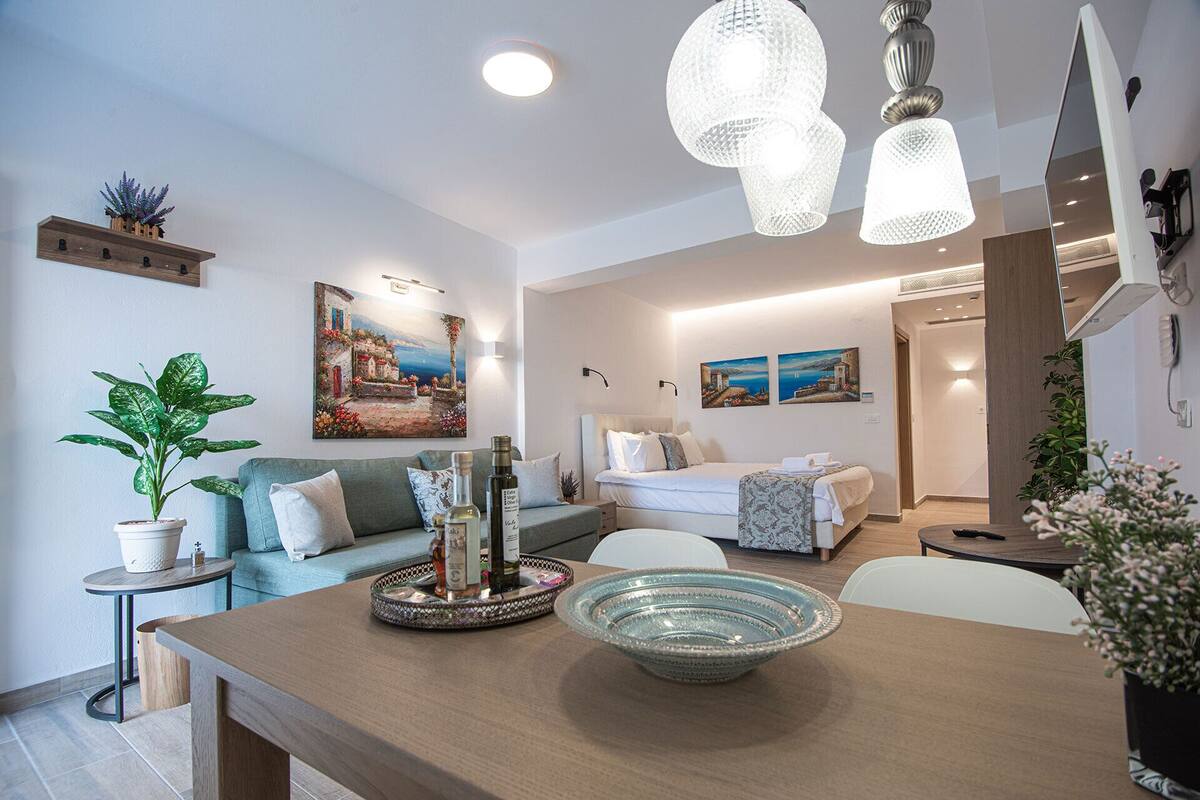 A modern living room with a wooden coffee table, a dining table, and a sofa. There are two paintings on the wall, a potted plant, and a chandelier.