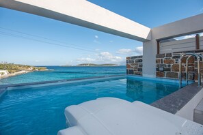 Une piscine avec vue sur la mer et les îles, des murs en pierre et un espace détente blanc.