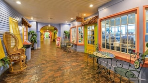 A corridor with brick flooring, wicker chairs, and tables, leading to a store with colorful walls and a window display.