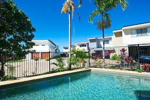 A swimming pool with a clear blue water, surrounded by a fence, palm trees, and modern buildings. - Magnetic Docks Townhouse 5 (Nelly Bay)