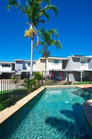 A swimming pool with clear water, surrounded by a fence, palm trees, and a row of modern buildings. - Magnetic Docks Townhouse 5 (Nelly Bay)