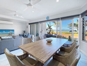 A modern dining area with a wooden table and wicker chairs, large windows with a view of the beach, and a ceiling fan. - Magnetic Docks Townhouse 5 (Nelly Bay)