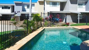 A swimming pool with clear water, surrounded by a fence, palm trees, and a row of modern buildings. - Magnetic Docks Townhouse 5 (Nelly Bay)