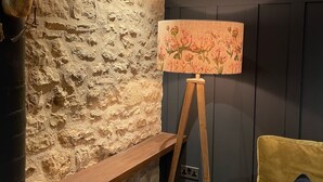 Quarto com parede de pedra, um cavalete de madeira com abajur floral, um banco de madeira e um sofá verde com almofada azul.