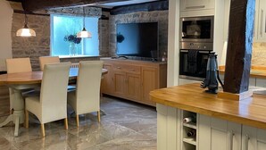 Cozinha moderna com parede de pedra, vigas de madeira, área de jantar com mesa e cadeiras, e forno embutido.