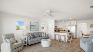 Ein geräumiges Wohnzimmer mit grauem Sofa, grauem Sessel und rundem Couchtisch. Im Hintergrund ist eine Küchenzeile mit Esstisch und Barhocker erkennbar.