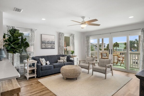 A spacious living room with a dark grey sofa, a round ottoman, and striped armchairs. A ceiling fan and a large window with curtains are visible. - Sand Buckets Unit A, Ocean View Beach Townhouse (Mexico Beach)