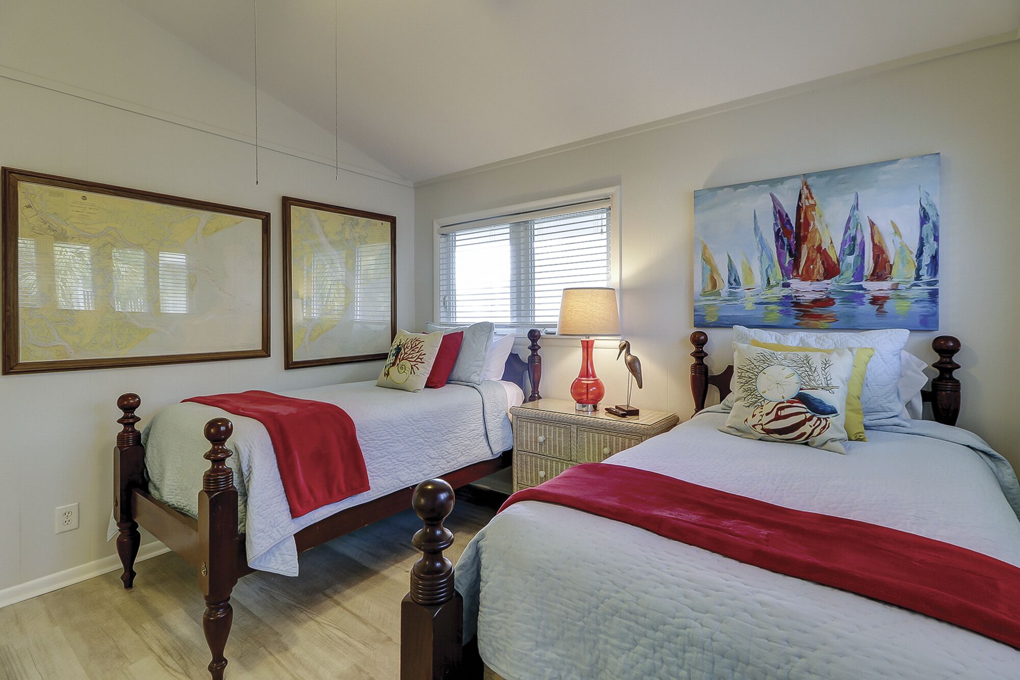 A hotel room with two beds, a window with blinds, a nightstand with a lamp, and artwork on the walls.