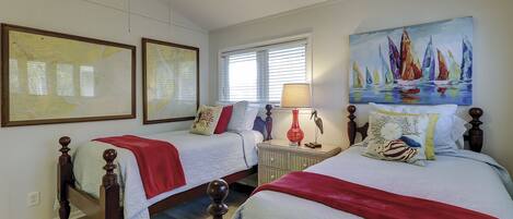 A hotel room with two beds, a window with blinds, a nightstand with a lamp, and artwork on the walls.