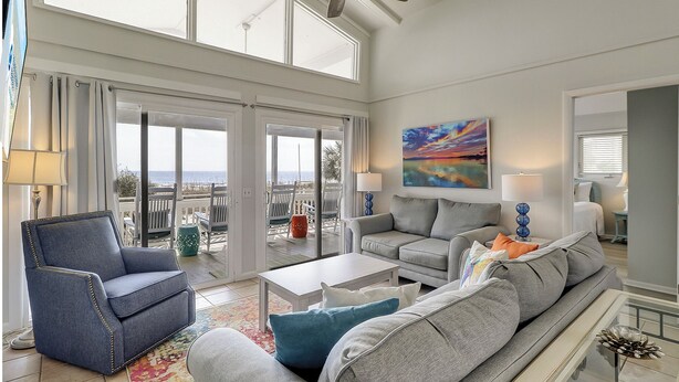 Een ruime woonkamer met een grote grijze bank, een blauwe fauteuil en een salontafel. Er is een balkon met uitzicht op zee en een plafondventilator.