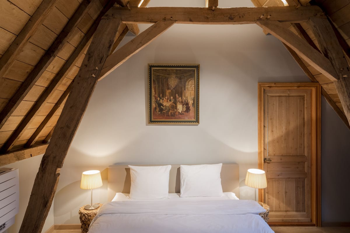 A bedroom with a wooden ceiling, a bed with white linens, two bedside lamps, and a wooden door.