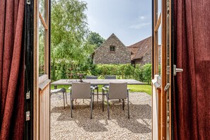 An open door leading to a patio with a table and chairs, a stone building in the background, and a view of a green landscape.