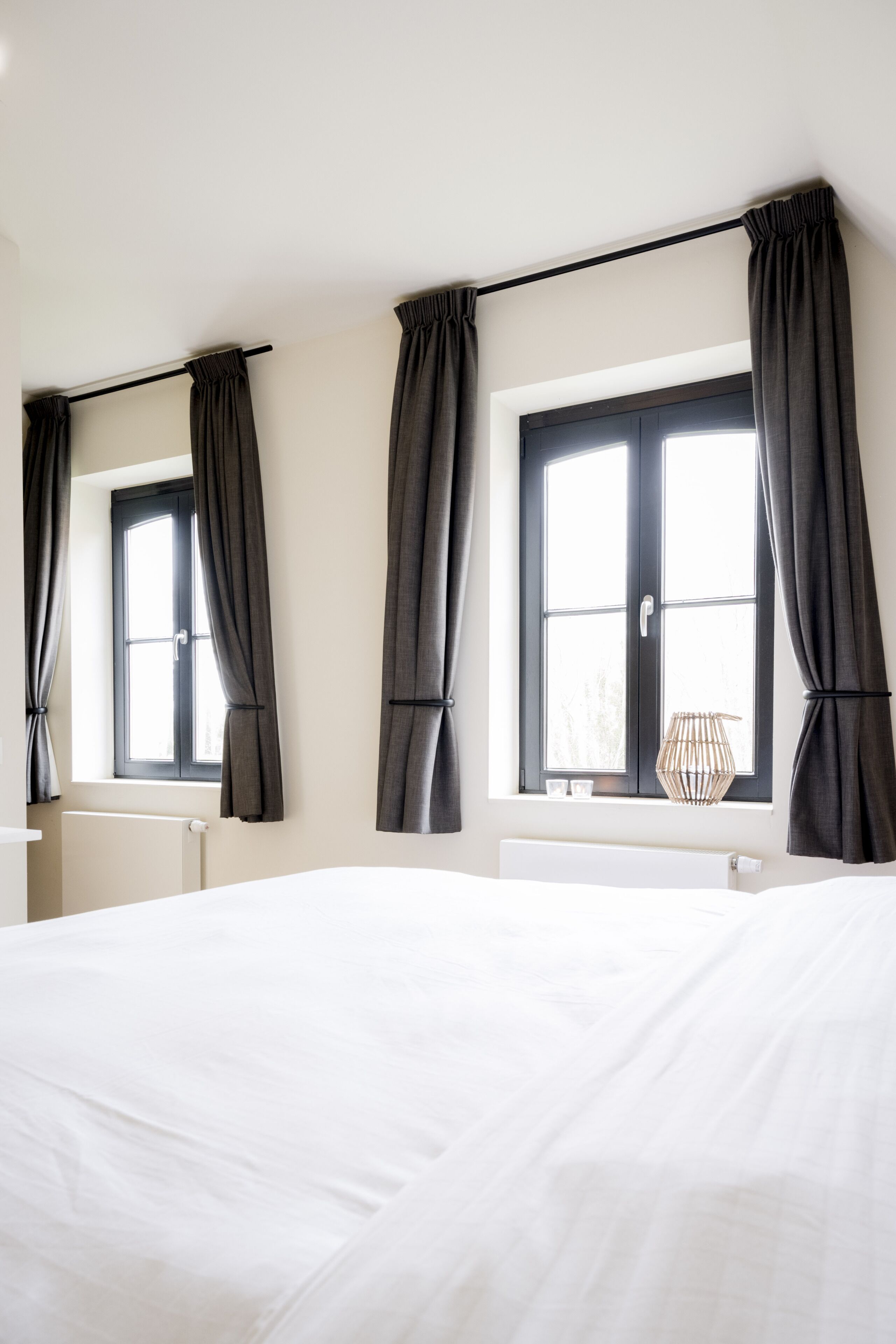 A bedroom with a large bed, two windows with black curtains, a radiator, and a small lamp on the windowsill.