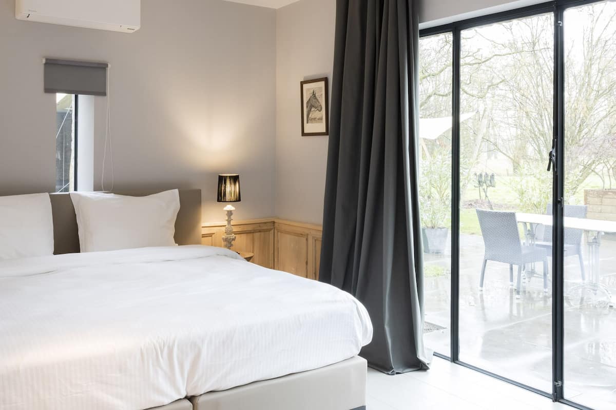 A modern bedroom with a large bed, a bedside table with a lamp, a window with blinds, and a view of the outdoors through a sliding glass door.