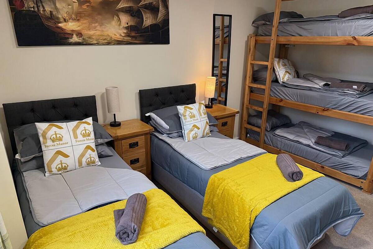A room with two beds, each with a headboard, a bedside table, and a lamp. There is a bunk bed with a ladder on the right side. A painting is on the wall above the beds.