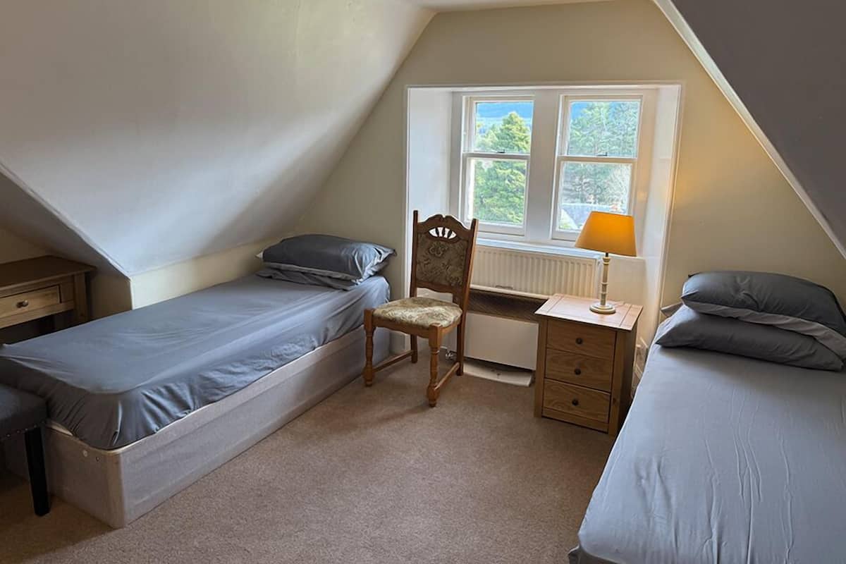 A small, triangular attic bedroom with two single beds, a desk with a chair, and a window with a view of greenery.