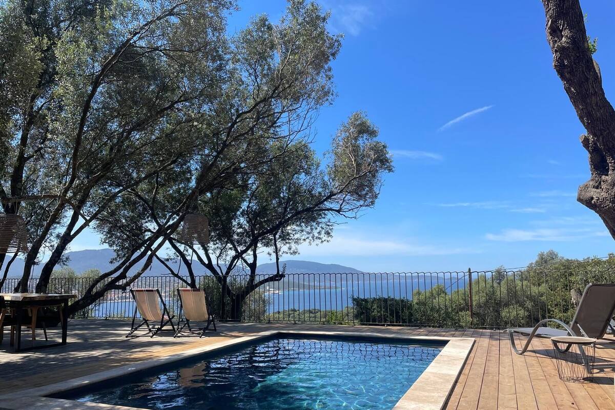 A rectangular pool with a wooden deck, surrounded by trees and a view of the sea.