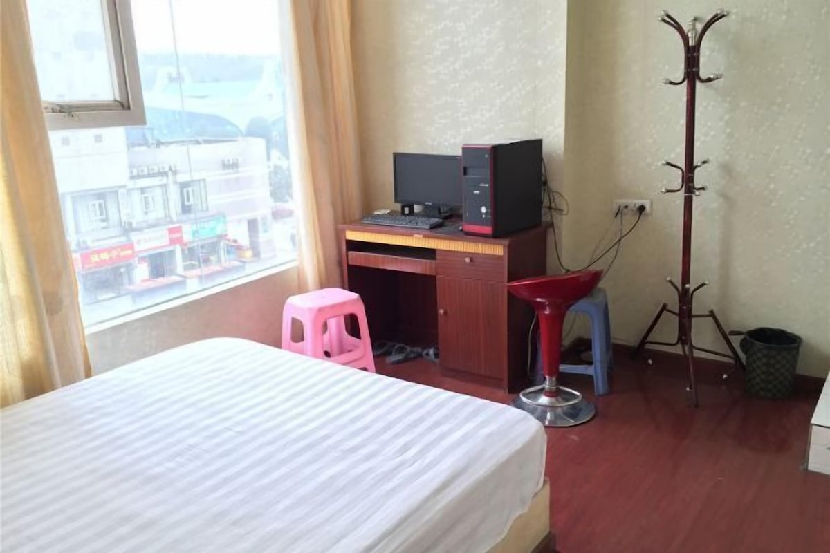 A room with a bed, a desk with a computer, a coat rack, and a window with a view of buildings.