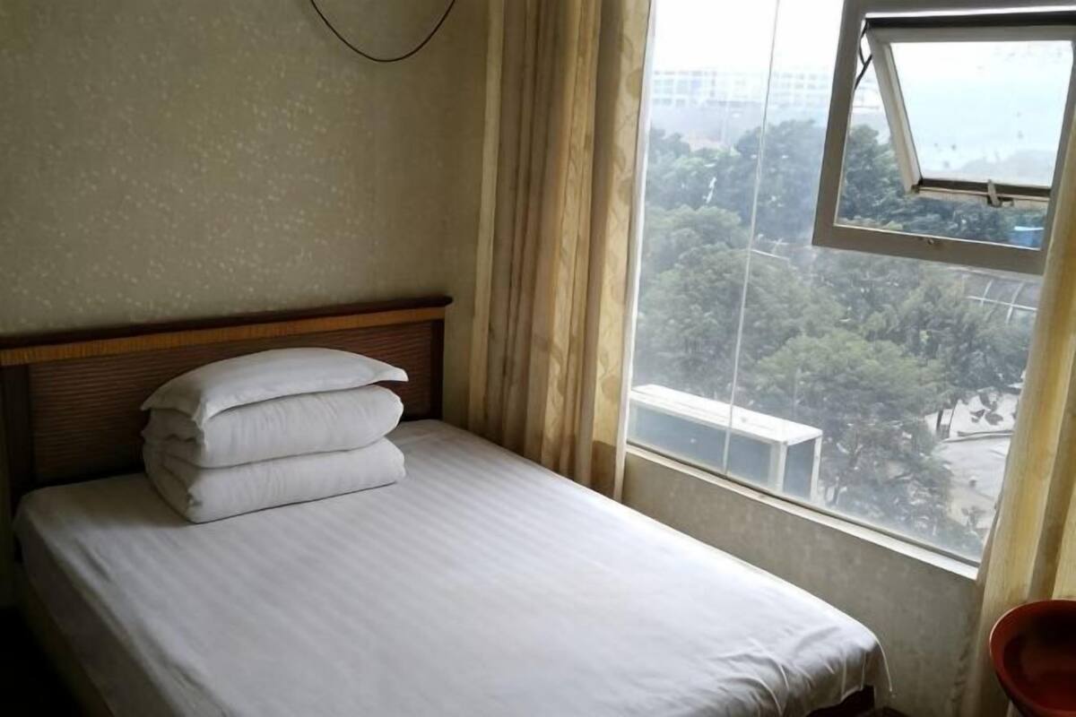 A single bed with white linens, a wooden headboard, and a window with a view of trees and buildings.