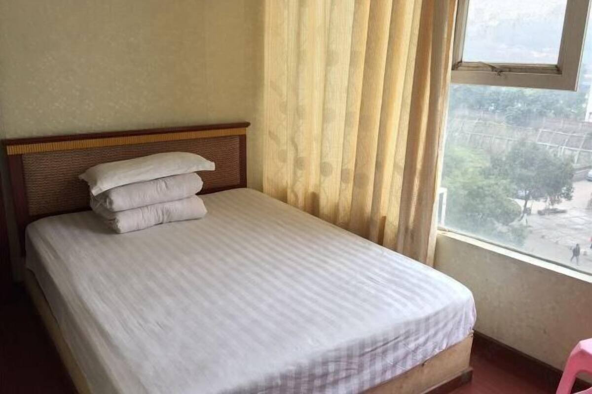 A single bed with a wooden headboard, a window with curtains, and a view of a street outside.