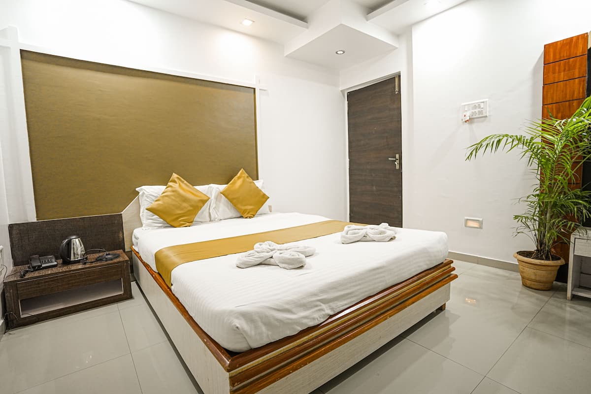 A modern hotel room with a large bed, a bedside table with a kettle, a potted plant, and a wooden headboard.