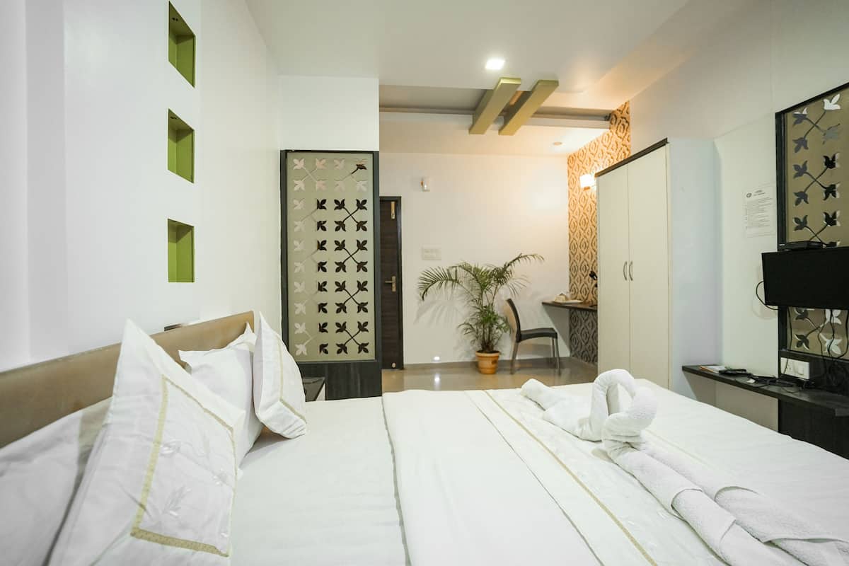 A hotel room with a neatly made bed, a desk with a chair, a potted plant, and a wardrobe.