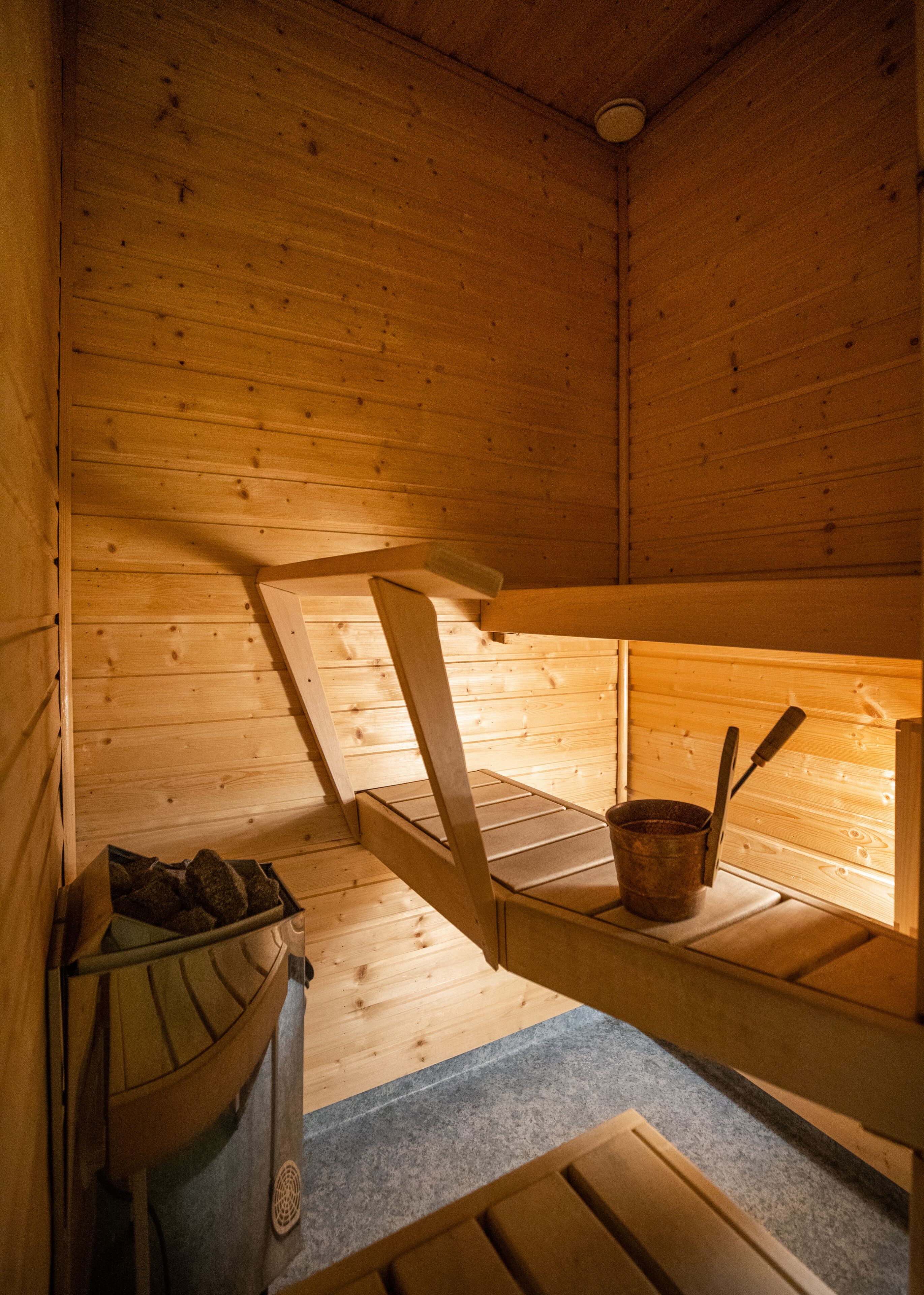 Interior d'una cabana de fusta amb un lliter, una taula petita amb un cub i un ganivet, i una caixa de guardat amb coses a dins.