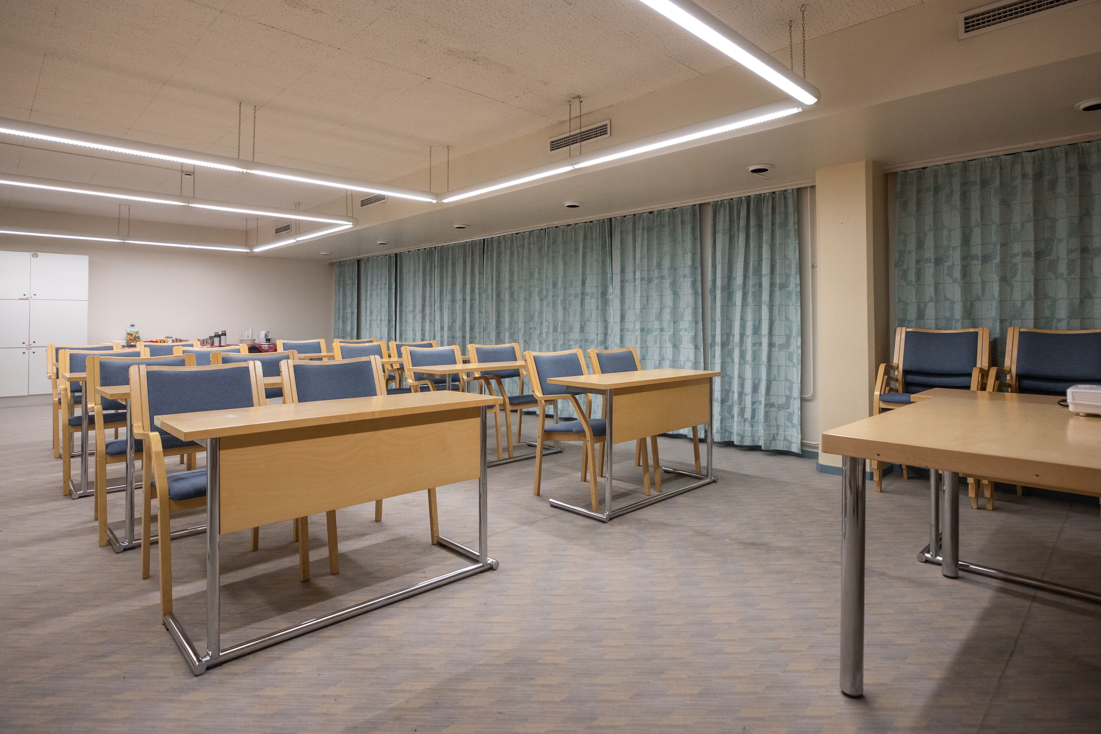 A conference room with rows of tables and chairs, a large curtain, and a plain ceiling with lighting fixtures.